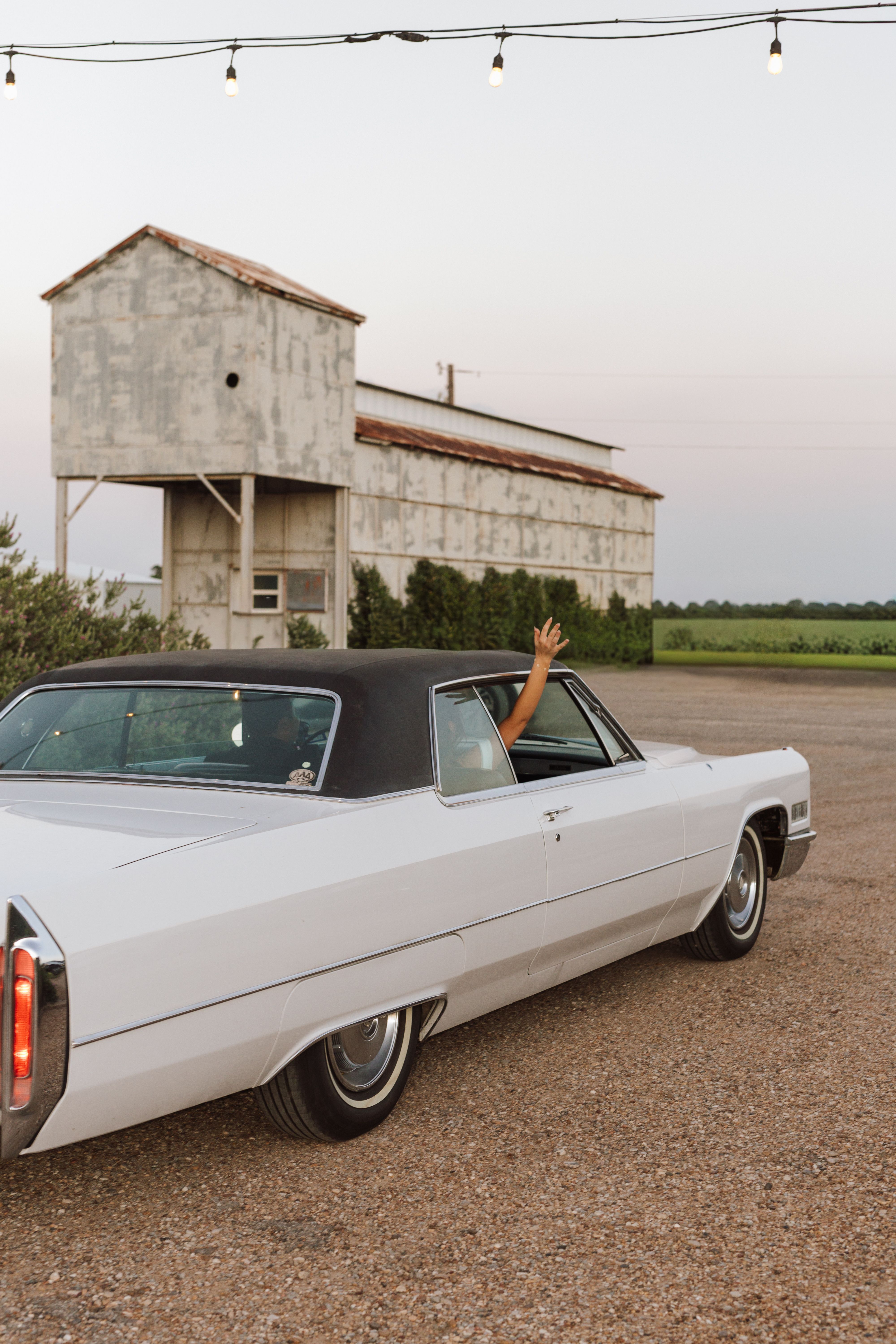 A bride waves out the window of a vintage white convertible as it pulls away from a rustic barn wedding venue at dusk near Montgomery, TX
